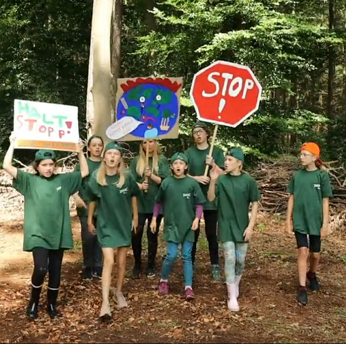 Es ist ein Photo abgebildet, auf welchem mehrere Kinder in grünen T-Shirts in einem Wald stehen. Drei Kinder halten Transparente hoch. Auf dem linken steht 